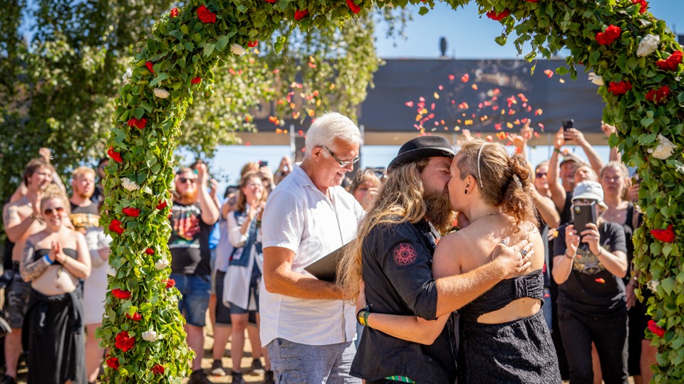 Ett nygift par kysser varandra, de står under en blomsterbåge på festivalområdet.